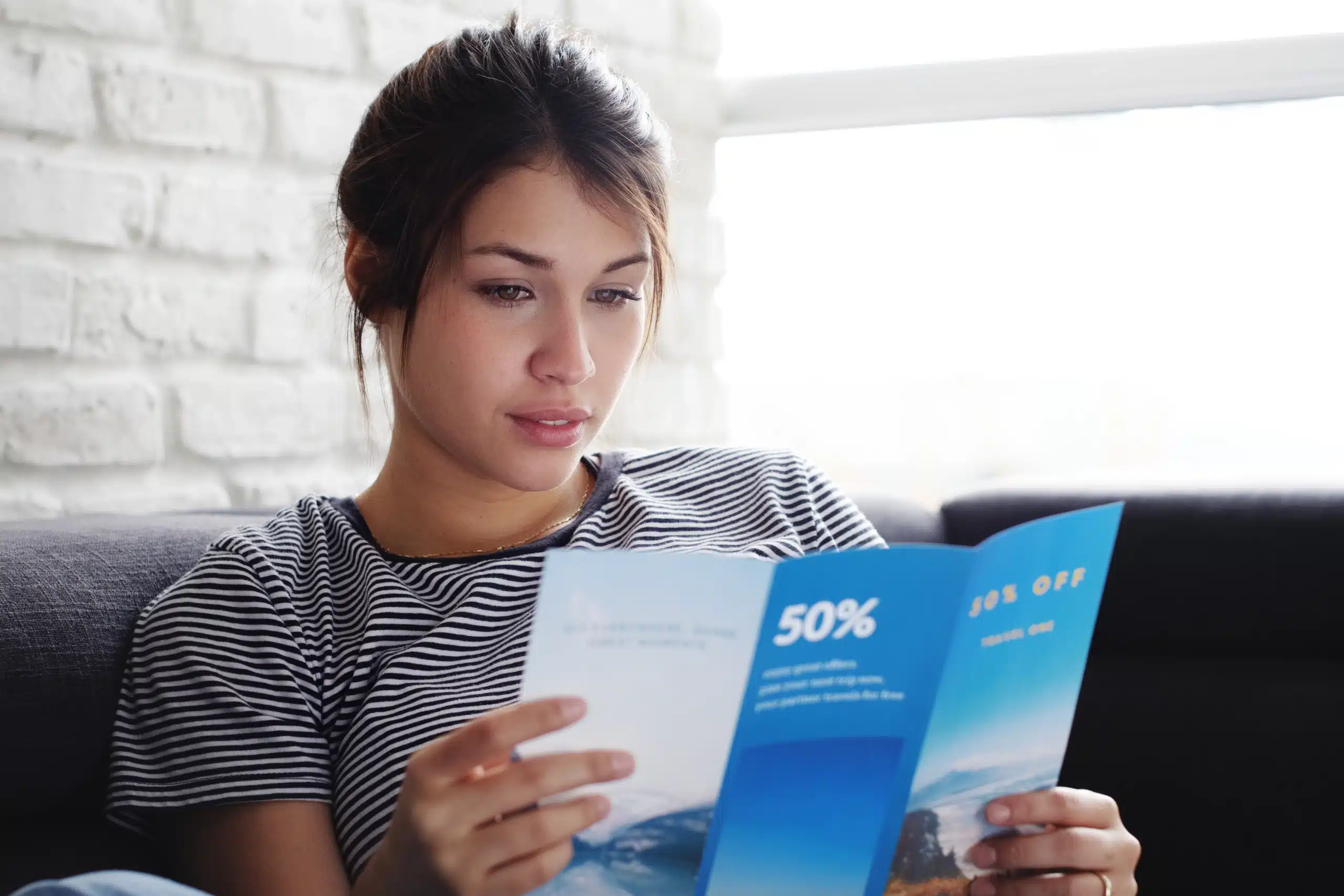 Young woman laying on couch at home, holding a travel flyer and planning next trip. Copy space on window
